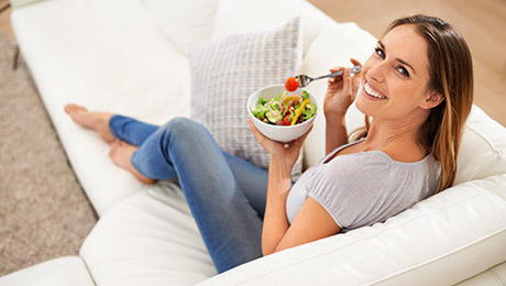 Frau mit Salat-Bowl sitzt auf Couch und lächelt in die Kamera