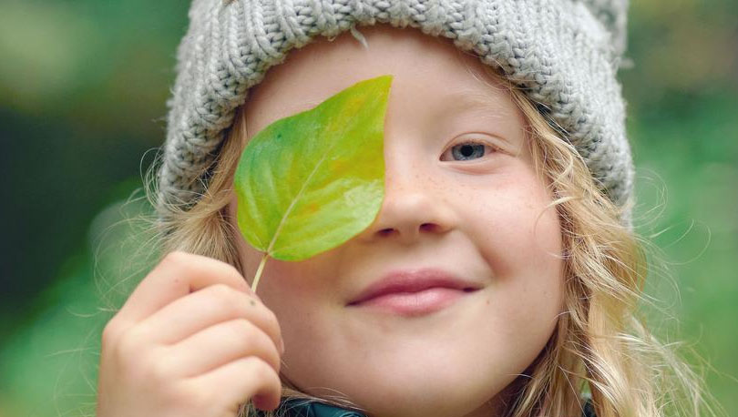 Kind mit Mütze hält sich grünes Blatt vor das Auge